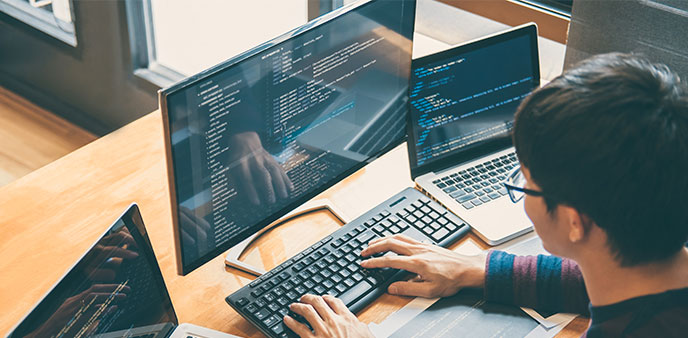 Person typing on keyboard at desk with multiple computer screens.